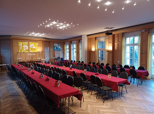 3 Lange, mit roten Tischdecken angerichtete Tischtafeln im großen Speisesaal mit schönen Deckenleuchten und hölzerner Fassade.