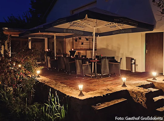 Außenansicht einer Terrasse mit einem Regenschirm unter welchem Stühle stehen, mit angenehmer indirekter Beleuchtung